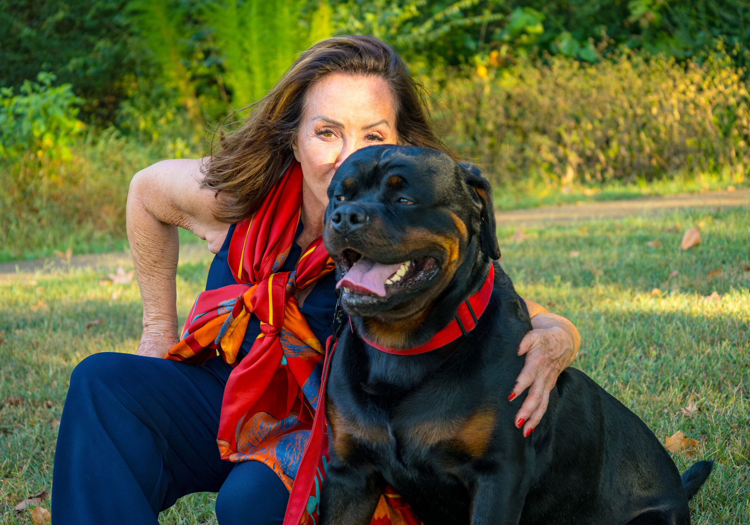 Woman sitting in the grass with her dog, surrounded by trees in the background