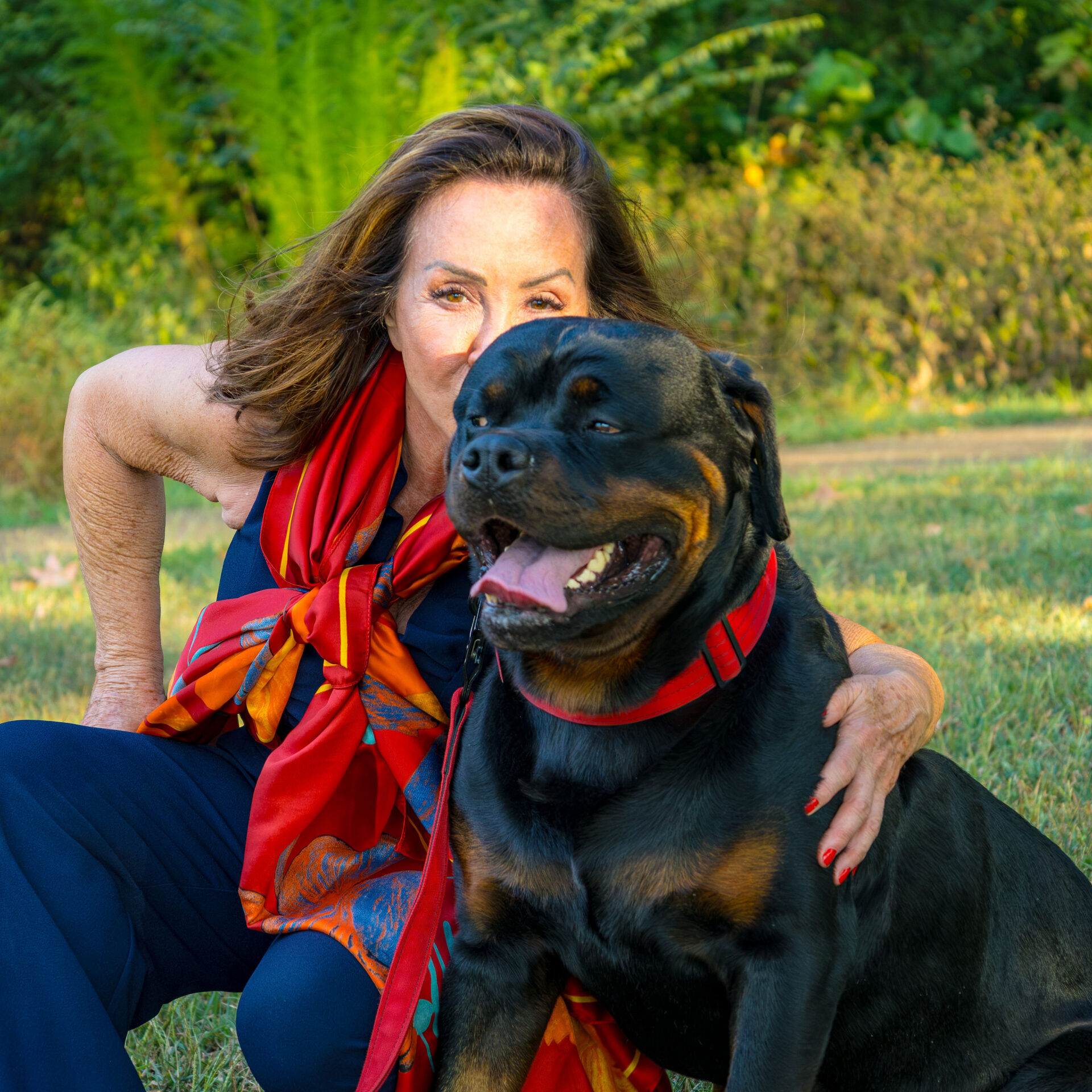 Woman sitting in the grass with her dog, surrounded by trees in the background