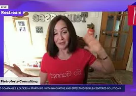 Woman in a shirt on a video call, with a wall behind her adorned with photo frames and text