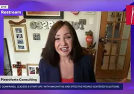 Woman in a jacket on a video call, with a wall behind her adorned with photo frames and other items